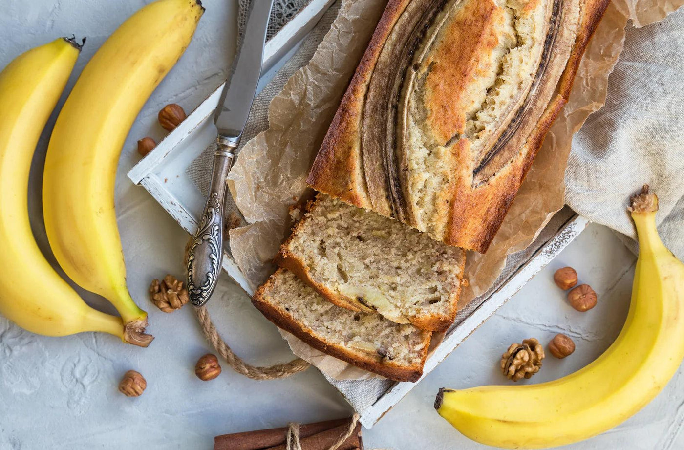 Banana bread maison avec deux tranches coupées, entouré de bananes mûres et de noix, posé sur une surface en papier parchemin avec un couteau ancien à côté.