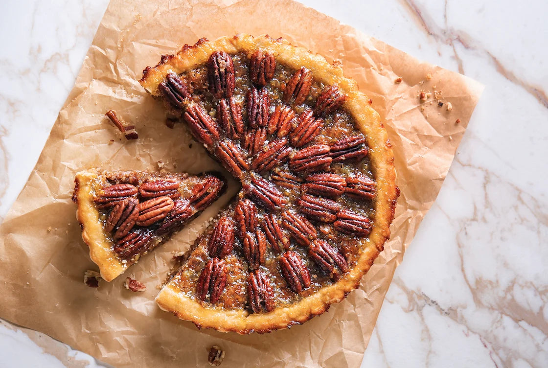Une tartelette au caramel et noix de pécan, garnie généreusement, reposant sur une assiette marron avec des noix de pécan entières en arrière-plan.