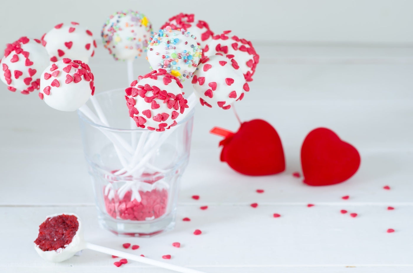 Des pop cakes enrobés de chocolat blanc et parsemés de cœurs en sucre rouges et multicolores, présentés dans un verre transparent comme support, sur un fond clair et épuré avec deux cœurs rouges en feutre.